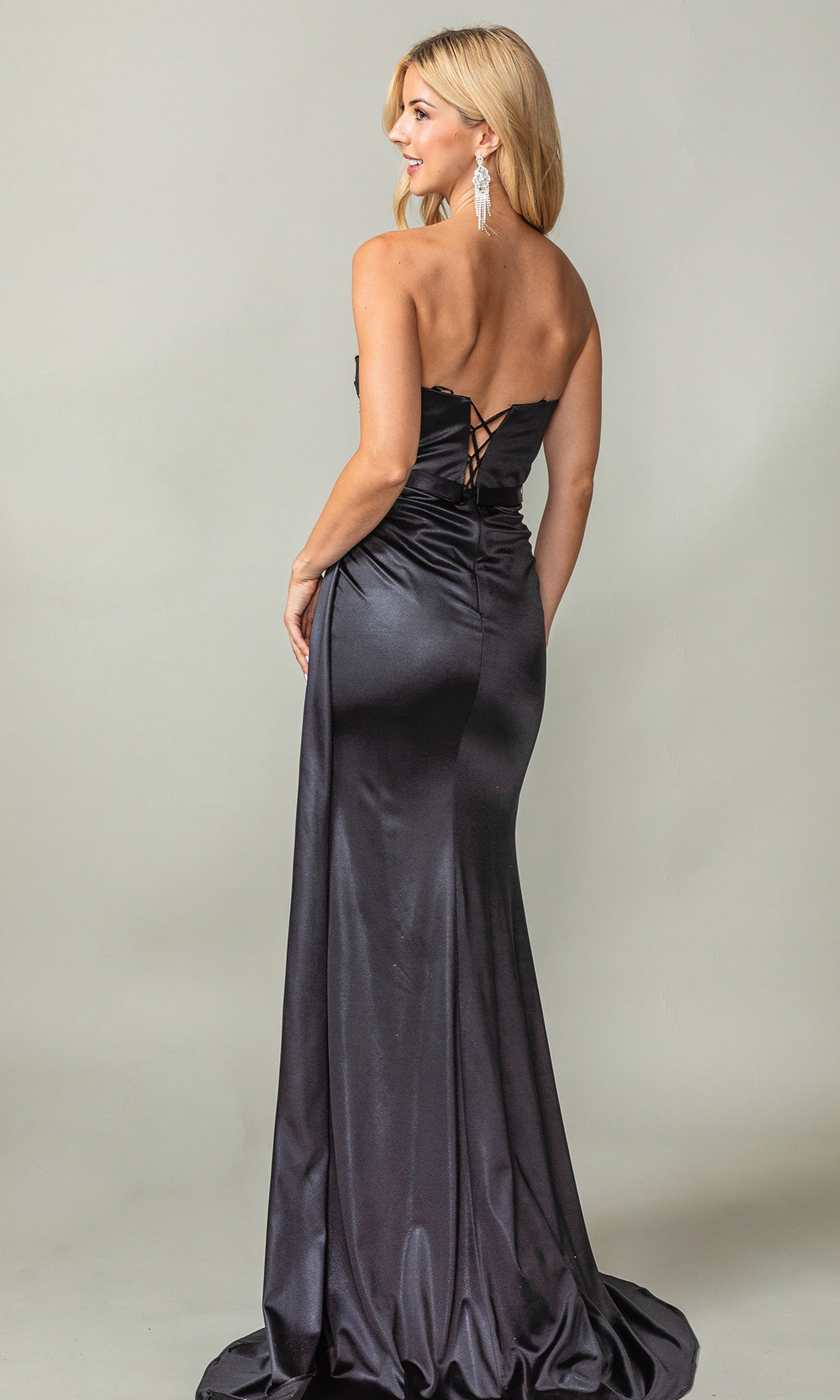 A woman in the Dancing Queen 4414 Strapless Long Sash Prom Dress, featuring a lace-up back and elegant satin finish, stands looking over her shoulder.